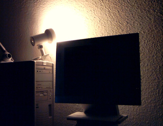 eine Leuchte, die auf einem PC-Gehäuse steht, leuchtet schwach auf die raufasertapezierte Wand; außerdem ein Flachbildschirm