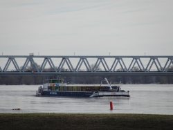 ein beladenes Containerbinnenschiff in der Elbe; im Hintergrund ein Teil der Eisenbahnbrücke über den Fluss