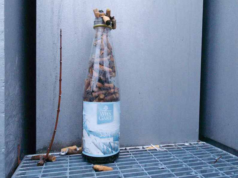 Eine Glasflasche für Saft von Wiesgart ist mit Zigarettenstummeln gefüllt. Zigarettenstummel liegen auch auf dem Gitterrost daneben.