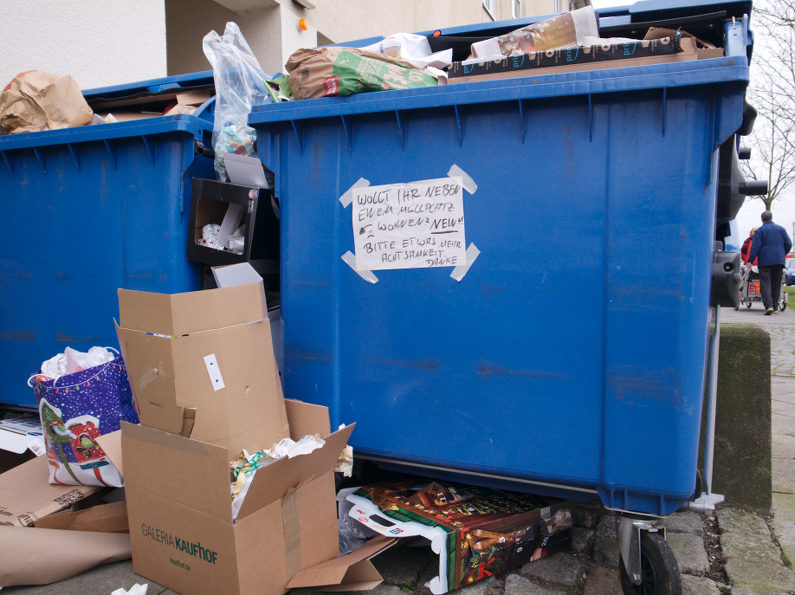 Pappe und Papier quillt aus blauen Containern. Ein handschriftlicher Hinweis bittet um Achtsamkeit; der Wohnort solle nicht zum Müllplatz werden.